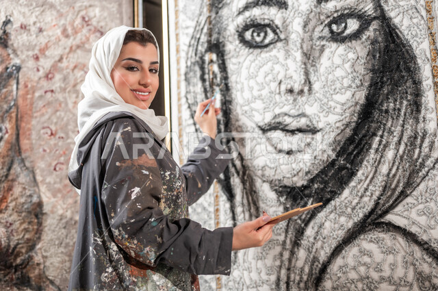 إنشاء تصاميم إبداعية فنية , رسامة عربية خليجية سعودية محجبة ترتدي مريلة الرسم تقوم بإتمام تفاصيل وجه الامرأة في اللوحات الجدارية المعلقة , معرض الرسومات و اللوحات التشكيلية في السعودية , النظر إلى الكاميرا بإيماءات السعادة و السرور