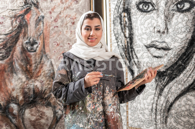 الوقوف أمام اللوحات الجدارية المعلقة , معرض الرسومات و اللوحات التشكيلية في السعودية , رسامة عربية خليجية سعودية محجبة ترتدي مريلة الرسم تحمل أدوات و معدات الرسم بيديها , الشغف بالفنون اليدوية