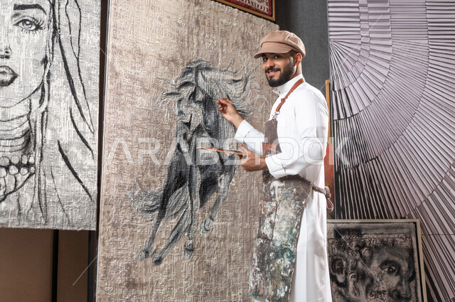 Exhibition of drawings and paintings in Saudi Arabia, passion for practicing manual arts and favorite hobbies, a Saudi Arabian Gulf painter wearing a traditional dress and a drawing apron completing the details of the horse, looking at the camera with gestures of happiness and pleasure