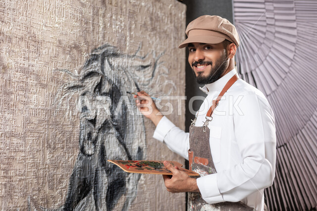 الشغف في ممارسة الفنون اليدوية و الهوايات المفضلة , صورة مقربة لرسام عربي خليجي سعودي يرتدي الثوب التقليدي و مريلة الرسم يقوم بإتمام تفاصيل الحصان , مفهوم الموهبة و الإبداع , النظر إلى الكاميرا بإيماءات السعادة و السرور