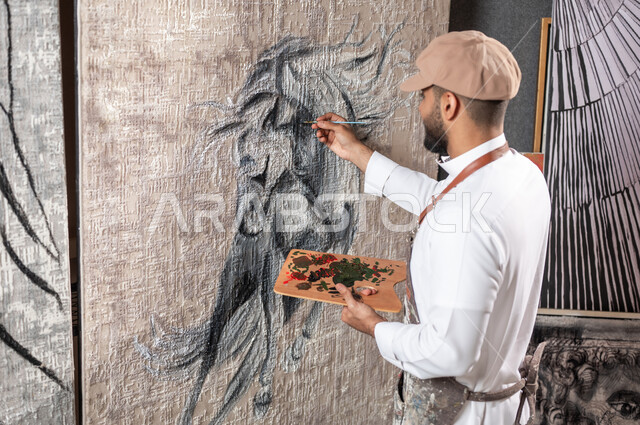 ممارسة هواية الفنون اليدوية , الشغف برسم الأحصنة , صورة مقربة لرسام عربي خليجي سعودي يرتدي الثوب التقليدي و مريلة الرسم  يقوم برسم حصان عربي أصيل , معرض الرسومات و اللوحات التشكيلية في السعودية , مفهوم الموهبة و الإبداع