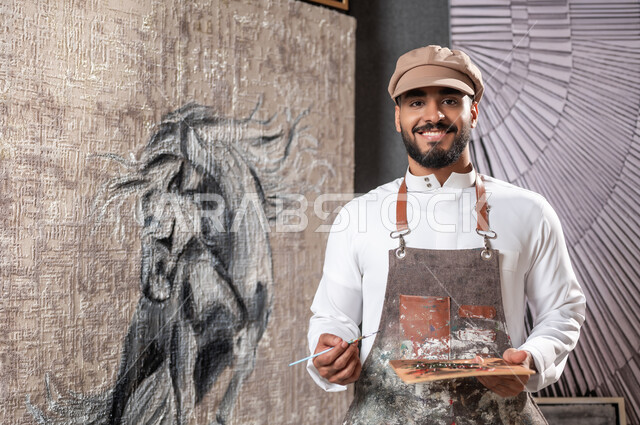 Passion for drawing horses, practicing the hobby of drawing, exhibition of artistic drawings and paintings, close-up picture of a Saudi Gulf Arab painter wearing a traditional dress and a drawing apron standing in front of a painting of a purebred Arabian horse, looking at the camera with gestures of happiness and pleasure