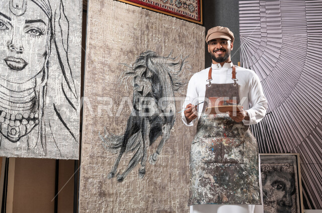 عرض المواهب و الإنجازات , ورشة عمل فنية , الشغف في ممارسة الفنون اليدوية و الهوايات المفضلة , رسام عربي خليجي سعودي يرتدي الثوب التقليدي و مريلة الرسم يحمل أدوات و معدات التلوين يقف أمام لوحة لحصان عربي أصيل , النظر إلى الكاميرا بإيماءات السعادة و السرور