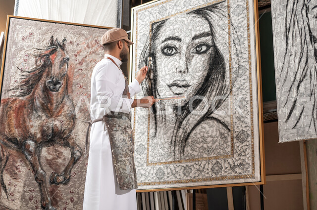 مفهوم الموهبة و الإبداع , الشغف في  ممارسة الفنون اليدوية و الهوايات المفضلة , رسام عربي خليجي سعودي يرتدي الثوب التقليدي و مريلة الرسم يقوم بإتمام تفاصيل وجه الامرأة في اللوحات الجدارية المعلقة  , معرض الرسومات و اللوحات التشكيلية في السعودية