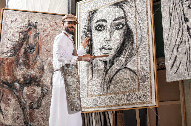 الشغف بالفنون اليدوية , ممارسة هواية الفن التشكيلي في ورشة عمل فنية , رسام عربي خليجي سعودي يرتدي الثوب التقليدي و مريلة الرسم يحمل أدوات و معدات التلوين , النظر إلى الكاميرا بإيماءات الفرح و السرور , معرض الرسومات و اللوحات التشكيلية في السعودية