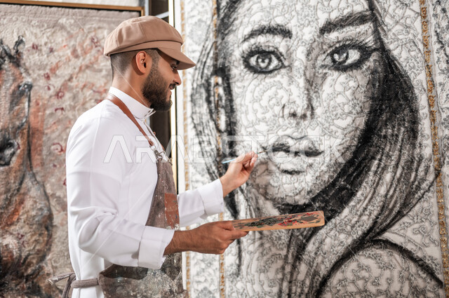 ورشة عمل فنية, ممارسة هواية الفن التشكيلي , الشغف بالفنون اليدوية , صورة مقربة لرسام عربي خليجي سعودي يرتدي الثوب التقليدي و مريلة الرسم يحمل أدوات و معدات التلوين , مفهوم الموهبة و الإبداع , معرض الرسومات و اللوحات التشكيلية في السعودية