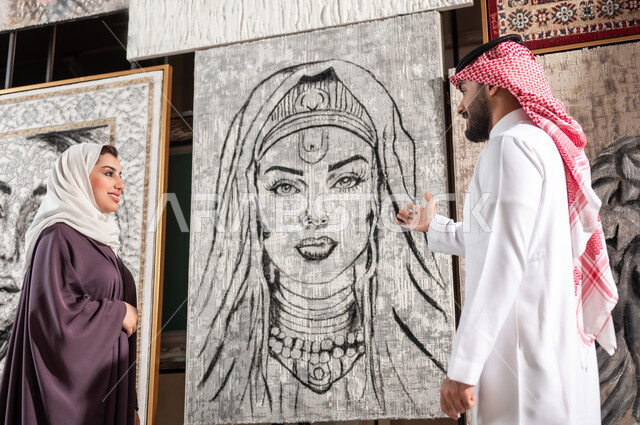 Passion for the talent of drawing, interest in handmade arts, exhibition of drawings and paintings, a Saudi Gulf Arab couple standing in front of artistic paintings, a man pointing with his hand to a painting of a woman in black and white