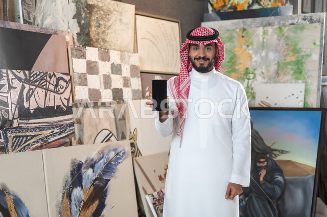 استخدام الأجهزة الحديثة و المتطورة , معرض الرسومات و اللوحات التشكيلية الفنية , رجل عربي خليجي سعودي يرتدي الثوب التقليدي و الشماغ يمسك جهاز الهاتف المحمول بيده , الاهتمام بالمواهب و الفنون اليدوية , عرض شاشة سوداء فارغة عبر الجوال