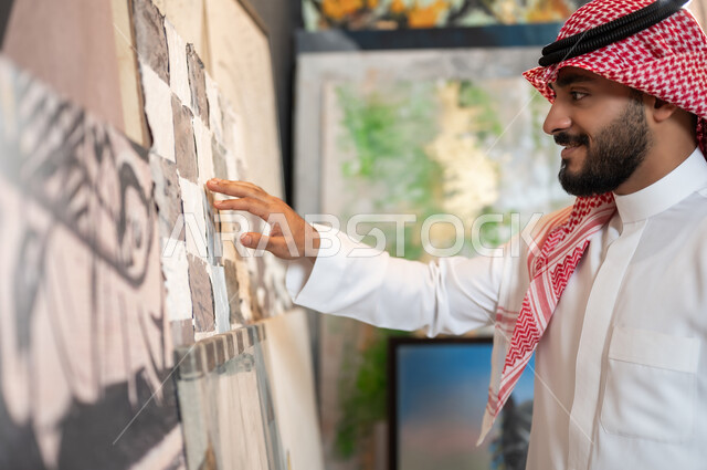 تأمل اللوحات و التصاميم الإبداعية , الشغف بالفنون اليدوية , صورة مقربة من الجانب لرجل عربي خليجي سعودي يرتدي الثوب التقليدي و الشماغ يقوم بلمس أحد اللوحات , ورشة عمل فنية , معرض الرسومات الفنية التشكيلية في السعودية