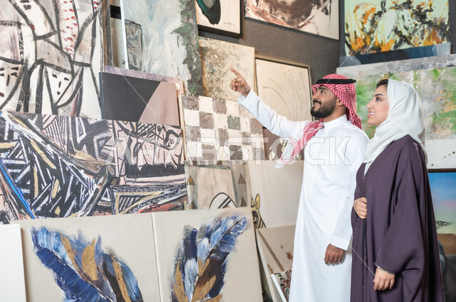 تصاميم فنية مميزة , أنشطة ترفيهية ممتعة , الشغف بالفن التشكيلي , زوجان عربيان خليجيان سعوديان يتجولان في معرض الرسومات و اللوحات , رجل يشير بإصبع السبابة إلى اللوحات الجدارية المعلقة