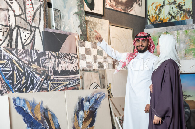 أنشطة ترفيهية ممتعة , الشغف بالفن التشكيلي , زوجان عربيان خليجيان سعوديان يتجولان في معرض الرسومات و اللوحات , رجل يشير بإصبع السبابة إلى اللوحات الجدارية المعلقة , تصاميم فنية مميزة