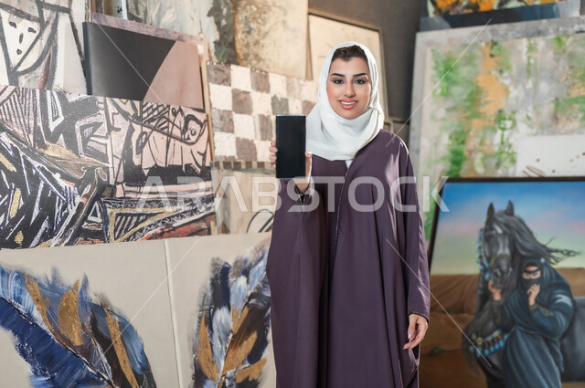 استخدام الأجهزة التقنية الحديثة , معرض الرسومات واللوحات التشكيلية الفنية , امرأة عربية خليجية سعودية محجبة ترتدي عباءة ملونة تمسك الهاتف المحمول بيدها , عرض شاشة سوداء فارغة عبر الجوال