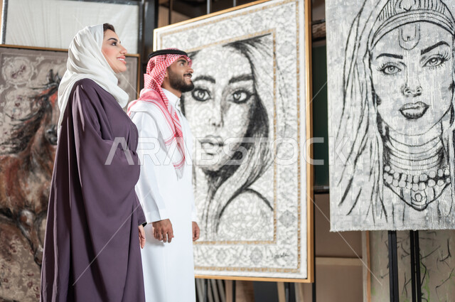 النظر إلى اللوحات الجدارية بإيماءات الفرح و السرور , صورة مقربة من الجانب لزوجان عربيان خليجيان سعوديان يتجولن في معرض الرسومات و الفنون التشكيلية يقومان بتأمل الرسوم , الاهتمام بالهوايات و المشاريع الفنية