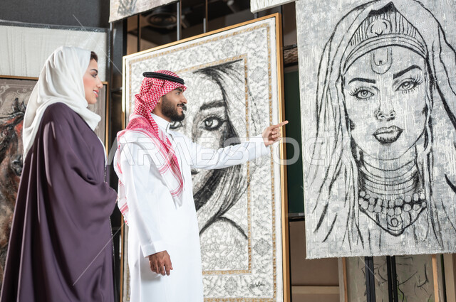 مفهوم المواهب و الفن , زوجان عربيان خليجيان سعوديان يتجولان في معرض الرسومات و الفنون التشكيلية , رجل يشير بإصبع السبابة إلى لوحة امرأة بالأبيض و الأسود , الاهتمام بالهوايات و المشاريع الفنية