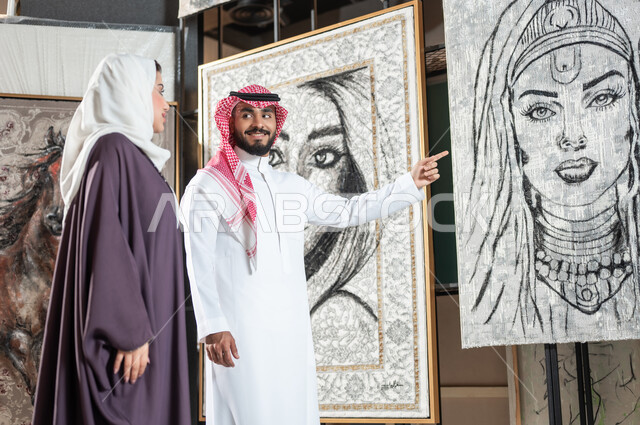 الاهتمام بالهوايات و المشاريع الفنية , زوجان عربيان خليجيان سعوديان يتجولان في معرض الرسومات و الفنون التشكيلية , رجل يشير بإصبع السبابة إلى لوحة امرأة بالأبيض و الأسود , مفهوم المواهب و الفن