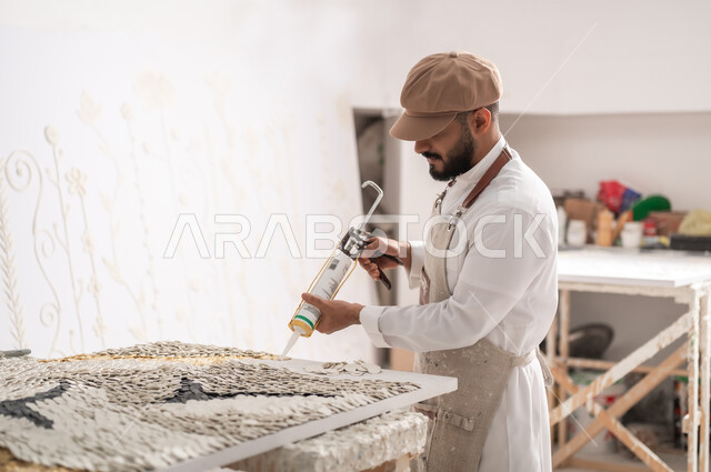 الشغف بالفنون اليدوية , استخدام الأدوات المناسبة لإنشاء لوحة مميزة , ممارسة هواية الرسم الجداري ثلاثي الأبعاد , رسام عربي خليجي سعودي يرتدي الثوب التقليدي و مريلة الرسم يقوم بتثبيت القطع البيضاء الصغيرة باستخدام فرد السيليكون , ورشة عمل فنية