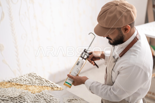 استخدام الأدوات المناسبة لإنشاء لوحة مميزة , ممارسة هواية فن التشكيل و الرسم بالجبس , صورة مقربة من الجانب لرسام عربي خليجي سعودي يرتدي الثوب التقليدي و مريلة الرسم يقوم بتثبيت القطع البيضاء الصغيرة باستخدام فرد السيليكون , الشغف بالفنون اليدوية , ورشة عمل فنية
