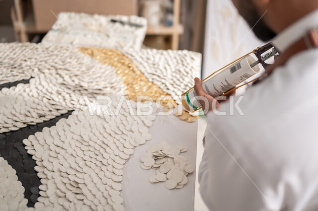 الشغف بالفنون اليدوية , ورشة عمل فنية , ممارسة هواية الرسم الجداري ثلاثي الأبعاد , صورة مقربة لرسام عربي خليجي سعودي يرتدي الثوب التقليدي و مريلة الرسم يقوم بتثبيت القطع البيضاء الصغيرة باستخدام فرد السيليكون