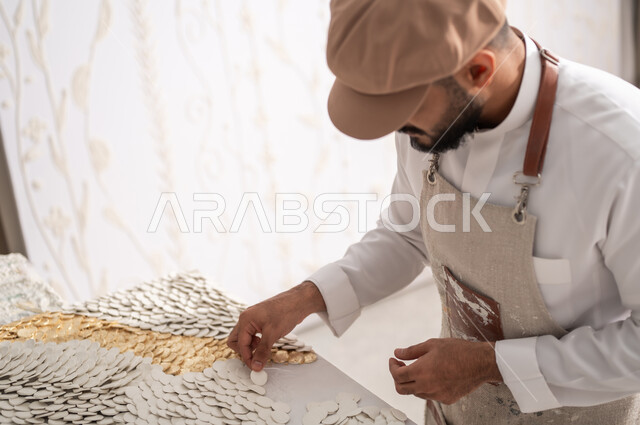 ممارسة هواية الرسم الجداري ثلاثي الأبعاد , صورة مقربة  لرسام عربي خليجي سعودي يرتدي الثوب التقليدي و مريلة الرسم يقوم بتثبيت القطع البيضاء الصغيرة , استخدام المواد المختلفة لإنشاء التصاميم , ورشة عمل فنية
