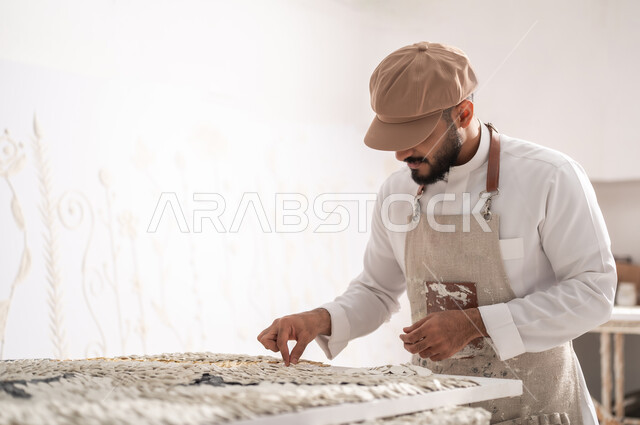 إنشاء تصاميم مميزة , ورشة عمل فنية , ممارسة هواية الرسم الجداري ثلاثي الأبعاد , رسام عربي خليجي سعودي يرتدي الثوب التقليدي و مريلة الرسم يقوم بتثبيت القطع البيضاء الصغيرة , إنجاز مشروع فني ناجح