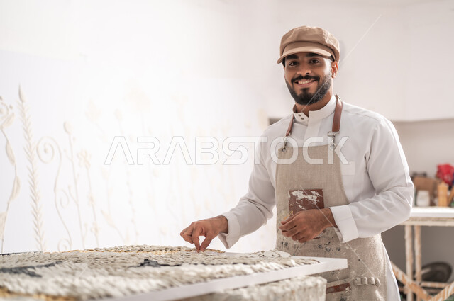 ممارسة هواية الرسم الجداري ثلاثي الأبعاد , استخدام قطع السيراميك لإنشاء تصاميم مميزة , صورة مقربة لرسام عربي خليجي سعودي مبتسم يرتدي الثوب التقليدي و مريلة الرسم يقوم بتثبيت القطع البيضاء الصغيرة في ورشة عمل فنية , النظر إلى الكاميرا بإيماءات الفرح و السرور , فن التشكيل و الرسم بالجبس