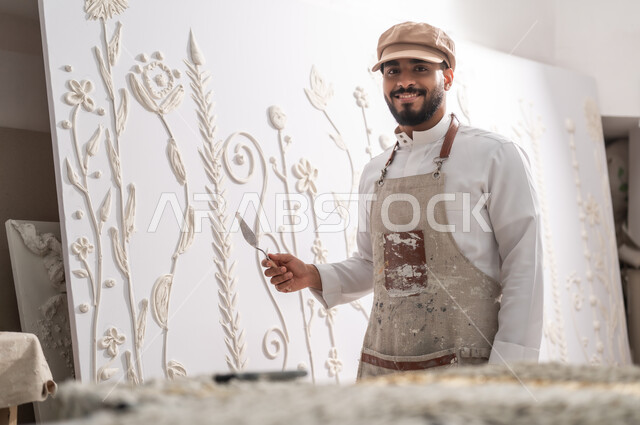 إنشاء تصاميم إبداعية بالجبس الأبيض , استخدام الأدوات المناسبة لإنجاز مشروع فني ناجح , ممارسة هواية الرسم الجداري ثلاثي الأبعاد , رسام عربي خليجي سعودي يرتدي الثوب التقليدي و مريلة الرسم ينظر إلى الكاميرا بإيماءات السعادة و السرور , ورشة عمل فنية