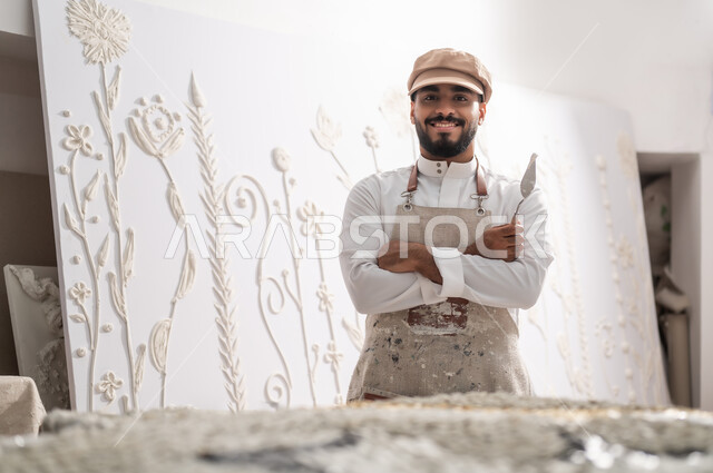 استخدام الأدوات المناسبة لإنشاء تصاميم بارزة على الجدران , النظر إلى الكاميرا بإيماءات الفرح و السرور , رسام عربي خليجي سعودي يرتدي الثوب التقليدي و مريلة الرسم , إنجاز مشروع فني ناجح , ورشة عمل فنية