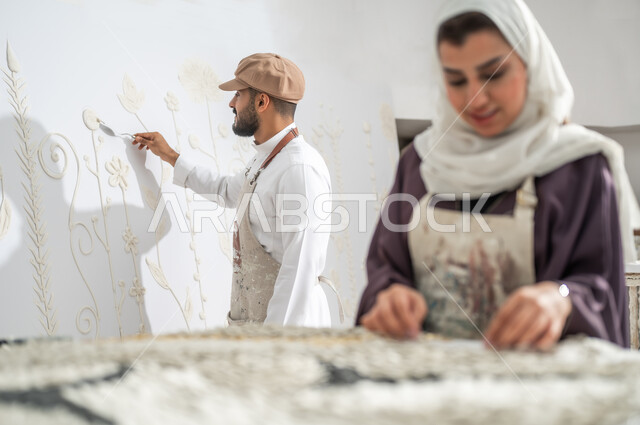 The painter installs small white pieces on the canvas, practicing the hobby of 3D wall painting, two Saudi Arabian Gulf painters wearing the traditional dress and a drawing apron, using ceramics to create designs, an art workshop