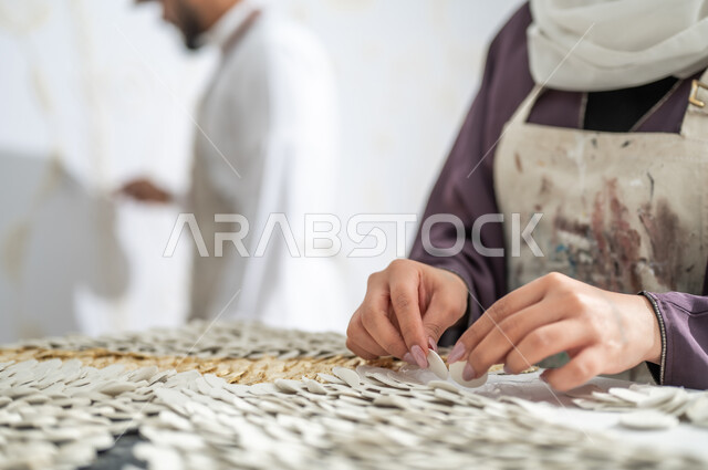 الفن الجداري بالجبس الأبيض ثلاثي الأبعاد استخدام قطع السيراميك لإنشاء التصاميم , صورة مقربة ليد رسامة عربية خليجية ترتدي عباءة ملونة مع مريلة الرسم تقوم بتثبيت القطع البيضاء الصغيرة , التعاون على إنجاز مشروع فني ناجح , ممارسة هواية الفنون اليدوية في ورشة عمل فنية