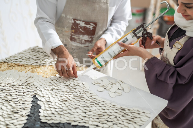 Three-dimensional wall art, cooperation to complete a successful art project, a close-up picture of a veiled Gulf artist using a silicone straightener standing with a Saudi Arab painter wearing a traditional dress and a drawing apron installing small white pieces, practicing the hobby of handmade arts in an art workshop
