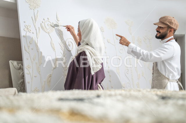 Fine art with white plaster, cooperation to complete a successful art project, directing gestures from a man to a woman, a close-up picture from the back of a Saudi Gulf Arab painter wearing a traditional dress and a drawing apron, pointing with his index finger to the painting, standing with a veiled painter, practicing the hobby of handmade arts in an art workshop
