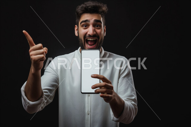 النظر الى الكاميرا بإيماءات الفرح والسرور، بورتريه مقرب لشاب عربي سعودي خليجي يرتدي الثوب التقليدي يمسك الهاتف المحمول بشاشة بيضاء فارغة يشير الى شيء ما, استخدام التقنيات والأجهزة الحديثة، خلفيه سوداء