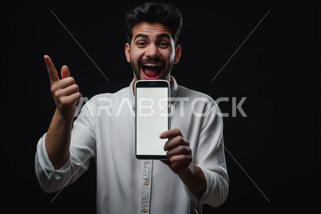 النظر الى الكاميرا بإيماءات الفرح والسرور، بورتريه مقرب لشاب عربي سعودي خليجي يرتدي الثوب التقليدي يمسك الهاتف المحمول بشاشة بيضاء فارغة يشير الى شيء ما, استخدام التقنيات والأجهزة الحديثة، خلفيه سوداء