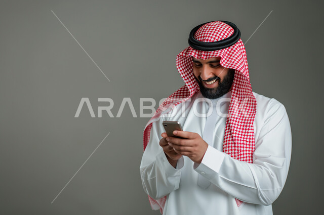 Chat and communicate with friends, browse social networking programs, a close-up portrait of a Saudi Gulf Arab young man wearing a shemagh and traditional dress, using a mobile phone with gestures of joy and pleasure, modern technologies and devices, gray background