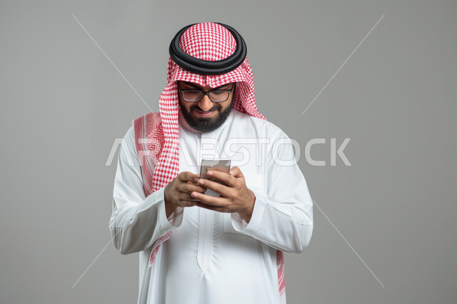 Chat and communicate with friends, browse social networking programs, a close-up portrait of a Saudi Gulf Arab young man wearing a shemagh and traditional dress, using a mobile phone with gestures of joy and pleasure, modern technologies and devices, gray background