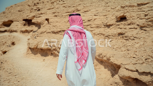 أماكن سياحية في المملكة، التكوينات والتشكيلات الصخرية الطبيعية في السعودية، صورة من الخلف لرجل عربي خليجي سعودي يرتدي الزي التقليدي يسير في الطرقات الصحراوية المتعرجة، منظر السماء الزرقاء
