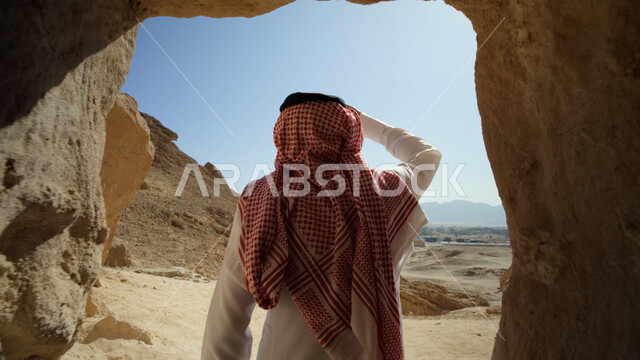 التشكيلات والتكوينات الصخرية في السعودية، الاستمتاع بالمناظر الطبيعية، رحلات السفاري في الصحراء، معالم وأماكن سياحية في المملكة، صورة مقربة من الخلف لرجل عربي خليجي سعودي يرتدي الزي التقليدي يقف داخل كهف صخري في المنطقة الصحراوية