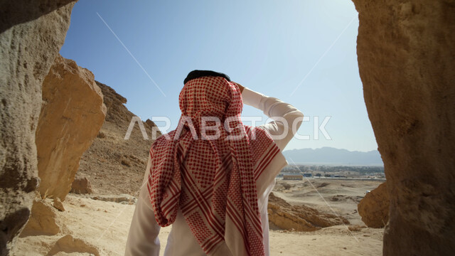 الاستمتاع بالمناظر الطبيعية، رحلات السفاري في الصحراء، التشكيلات والتكوينات الصخرية في السعودية، معالم وأماكن سياحية في المملكة، صورة مقربة من الخلف لرجل عربي خليجي سعودي يرتدي الزي التقليدي يقف داخل كهف صخري