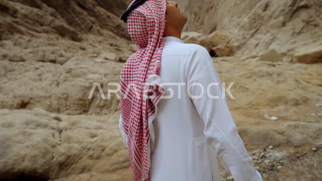 التشكيلات والتكوينات الصخرية الطبيعية في السعودية، معالم وأماكن سياحية في المملكة، صورة من الخلف لرجل عربي خليجي سعودي يرتدي الزي التقليدي يسير في طرقات المنطقة الصحراوية يقوم بالنظر إلى شيء ما