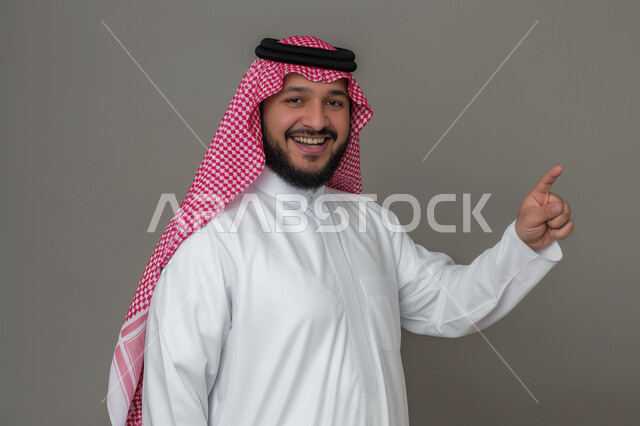 Concern with elegance and external appearance, a close-up portrait of a Saudi Gulf Arab young man wearing a shemagh and traditional dress, pointing with his index finger at something, looking at the camera with gestures of pleasure, the concept of masculinity and self-confidence, gray background