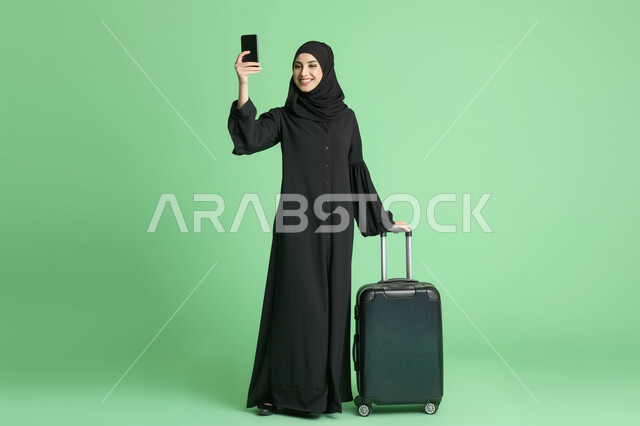 تأكيد الحجز الالكتروني عبر الجوال، بورتريه لشابة عربية خليجية سعودية محجبة ترتدي العباءة السوداء تمسك بيدها حقيبة السفر والهاتف المحمول، الاستعداد لبدء الرحلة في العطلة الصيفيه بحماس وسرور، صورة الجسم بالطول الكامل، خلفيه خضراء