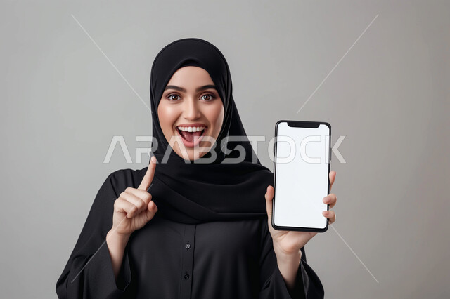 النظر الى الكاميرا بإيماءات الفرح والسرور، بورتريه مقرب لشابة عربية سعودية خليجية محجبة ترتدي العباءة السوداء تحمل بيدها الهاتف المحمول بشاشة بيضاء فارغة تشير بإصبع السبابة الى شيء ما, استخدام التقنيات والأجهزة الحديثة، خلفيه رمادية
