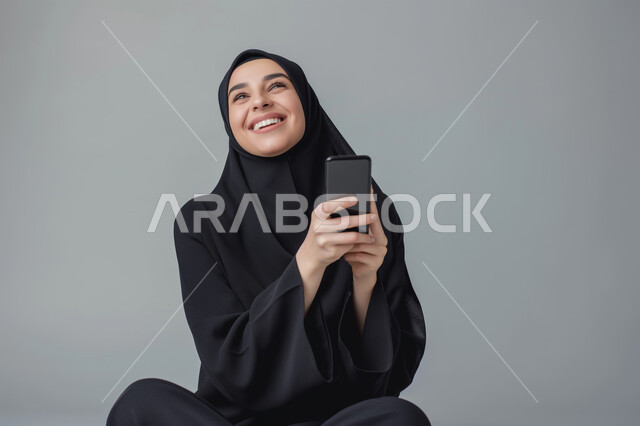 الدردشة والمحادثة مع الأصدقاء، تصفح برامج التواصل الاجتماعي، بورتريه مقرب لشابة عربية سعودية خليجية ترتدي العباءة السوداء التقليدية تقوم باستخدام الهاتف المحمول بإيماءات الفرح والسرور, التقنيات والأجهزة الحديثة، خلفيه رمادية