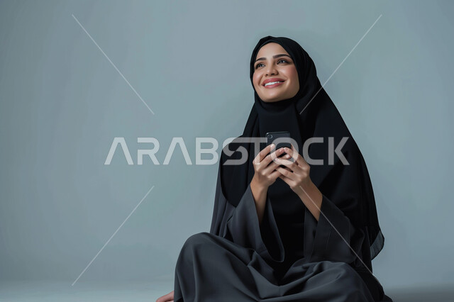 الدردشة والمحادثة مع الأصدقاء، تصفح برامج التواصل الاجتماعي، بورتريه مقرب لشابة عربية سعودية خليجية ترتدي العباءة السوداء التقليدية تقوم باستخدام الهاتف المحمول بإيماءات الفرح والسرور, التقنيات والأجهزة الحديثة، خلفيه رمادية