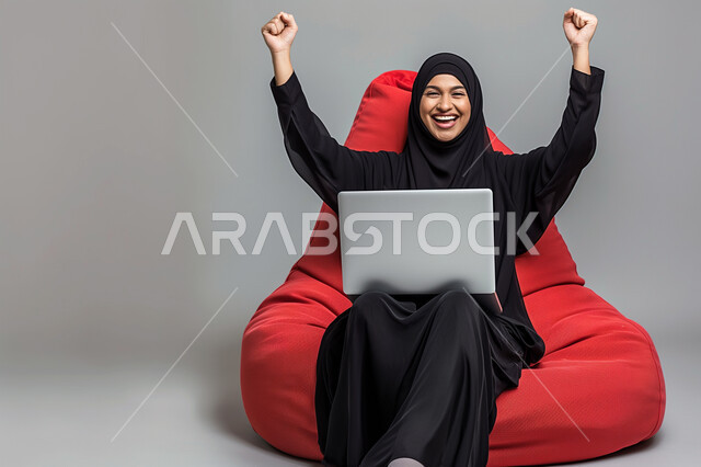 تعابير الفوز والانتصار، مفهوم النجاح وتحقيق الأهداف، بورتريه لشابة عربية سعودية خليجية محجبة ترتدي العباءة السوداء تجلس على كرسي مريح يقوم باستخدام جهاز الحاسوب المحمول ترفع يديها للاعلى بإيماءات الفرح والسرور, التقنيات والأجهزة الحديثة، خلفيه رمادية
