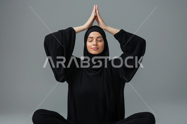 المحافظة على التوازن الجسدي، ايماءات الراحة والطمأنينة، أسلوب حياة صحي، بورتريه لشابة عربية خليجية سعودية محجبة ترتدي العباءة السوداء تقوم بممارسة تمارين اليوغا، مفهوم الرشاقة واللياقة، خلفية رمادية