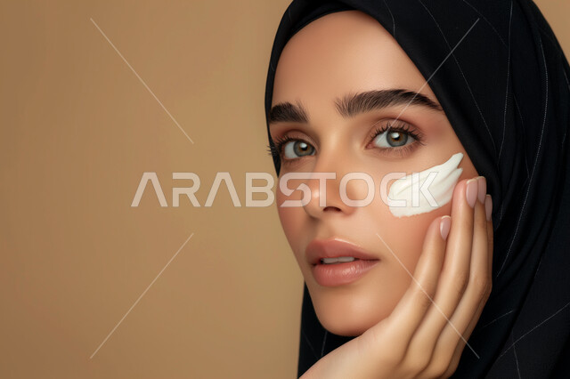 النظر إلى الكاميرا بإيماءات السرور، الاهتمام بالمظهر الخارجي، مفهوم العناية بنضارة وتحسين البشرة، بورتريه مقرب لامرأة عربية خليجية سعودية محجبة مبتسمة تضع كريم الترطيب، استخدام كريمات ومكونات تجميلية، خلفية ملونة