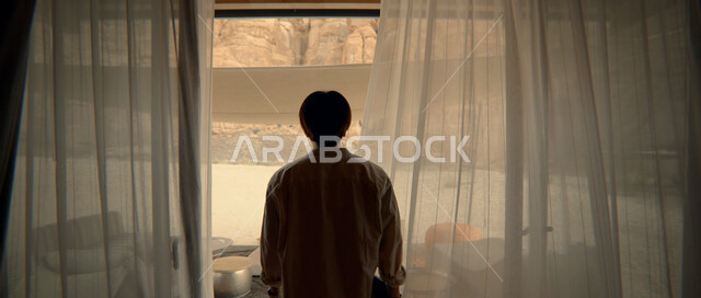 الاستمتاع بالنظر الى العالم الخارجي من خلال النافذة، التأمل وشرود الذهن، التفكير في شيء ما، صورة من الخلف لرجل عربي خليجي سعودي يقف امام واجهه زجاجية كبيرة وينظر الى الطبيعة الصخرية في أحد المنتجعات الراقية