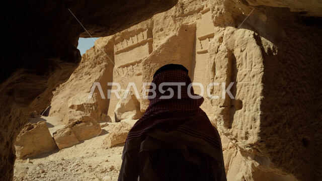 زيارة المناطق السياحية في صحراء المملكة العربية السعودية، التكوينات والتشكيلات الصخرية، صورة مقربة لرجل عربي خليجي سعودي يرتدي الزي التقليدي يقف داخل كهف يقوم بالنظر إلى المعالم التاريخية