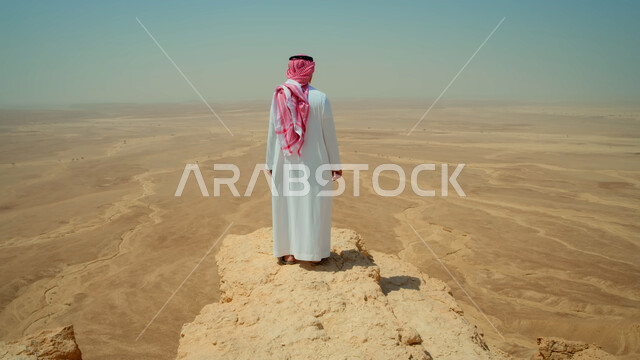 الأجواء الهادئة في صحراء المملكة العربية السعودية، صورة من الخلف لرجل عربي خليجي سعودي يرتدي الزي التقليدي يقف على قمة جبل صخري يقوم بتأمل المنطقة الصحراوية القاحلة، الاستمتاع بمشاهدة المناظر الطبعية